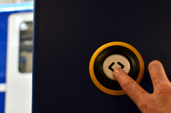 The hand presses the button for stop in the train. Orange door of train carriage with the electric blue button press to open sliding mechanical door at a train station platform. Small sharpness