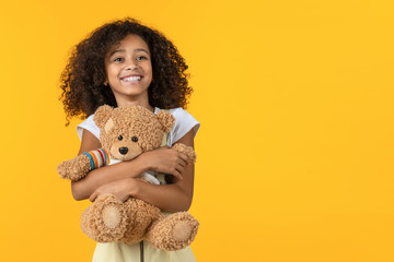 Little cute african girl hugging teddy bear toy isolated on yellow background