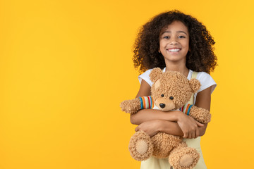 Portrait of smiling african girl hugging teddy bear toy isolated on yellow background