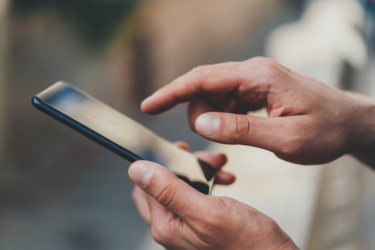 Closeup image of male hands using modern smartphone device outdoor, hipster man typing an sms message via cellphone while walking in urban streets, communication concept