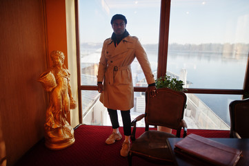 Handsome african american man posing  inside room with sunlight shadows in black hat and beige coat.