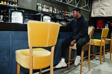 Handsome african american man posing  inside night club in black hat, sitting on bar counter.