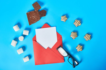 Scattered sweets, striped candies, cylindrical marshmallows, broken chocolate near glasses and envelope with white blank paper sheet lies on blue desk. Love letters concept. Top view. Close-up