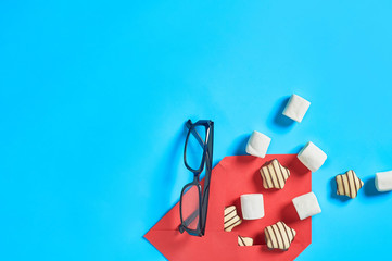 Red paper envelope with scattered sweets, striped candies, cylindrical marshmallows near optical glasses in black plastic frame lies on blue desk. Love letters concept. Space for text. Top view