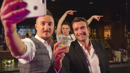 Two men stand at the bar and make a selfie with alcohol and bartender 
