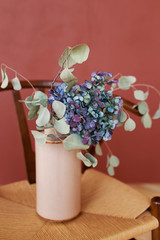 Blue and purple hydrangea in a beige vase stands on a wooden chair on a red background, eco-friendly home decoration, flowers for March 8
