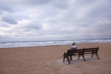 Fototapeta na wymiar Teenager sitting on a bench on an empty beach, cloudy day. The concept of loneliness.