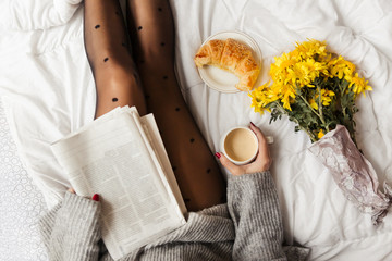 Woman sitting in bed reading newspaper and having breakfast. Top view 