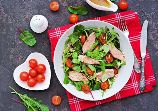 Healthy salad made from green salad mix with baked duck, stewed cherry tomatoes and pine nuts in a gray bowl on a dark brown concrete background. Healthy salads recipes.