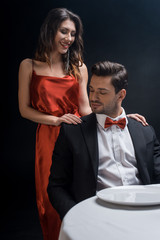 Smiling woman in elegant dress standing by handsome man at served table isolated on black