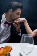 Side view of man in suit kissing hand of woman at served table on black background with smoke