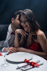 Handsome man whispering to beautiful smiling woman at served table on black background with smoke