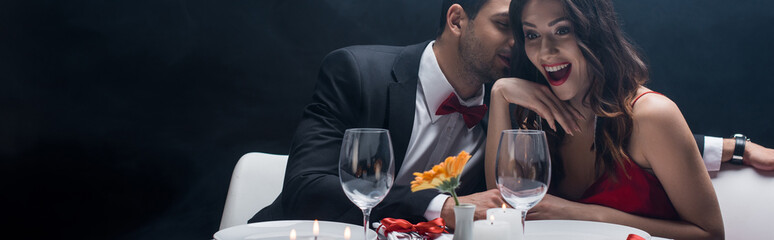 Panoramic shot of handsome man whispering to shocked woman during romantic dinner isolated on black