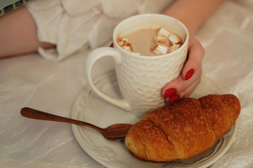 A young woman eats a croissant for Breakfast and drinks coffee. A woman's hand holds a mug of coffee on a plate is a croissant