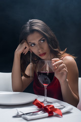 Attractive woman with hand by head touching wine glass at served table on black background with smoke
