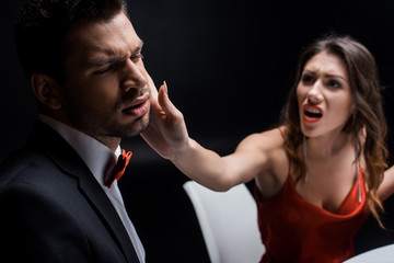 Selective focus of screaming woman slapping elegant boyfriend at table isolated on black