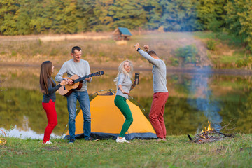 Group of happy friends with guitar, having fun outdoor, dancing and jumping near the lake in the park background the Beautiful sky. Camping fun