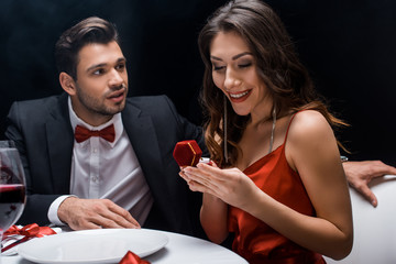 Handsome man looking at smiling girlfriend with jewelry ring during romantic dinner isolated on black