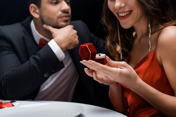 Selective focus of smiling woman holding jewelry ring in box by handsome boyfriend at served table isolated on black