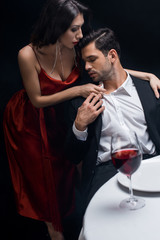 Selective focus of elegant woman unbuttoning shirt of handsome man by wine glass on table isolated on black