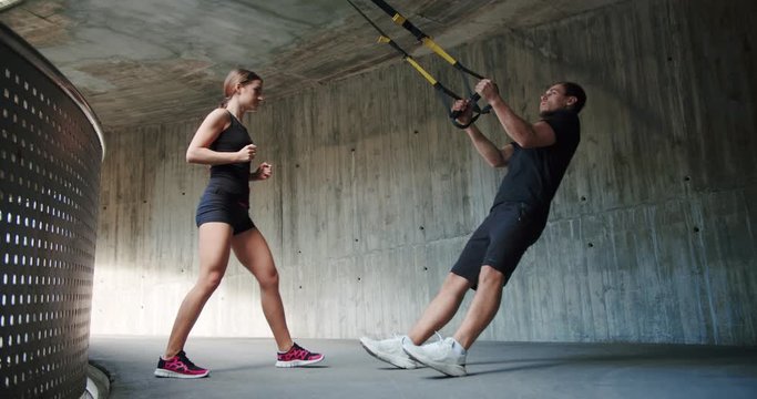 Strong Caucasian female personal trainer explaining pull ups exercise to motivated guy, effort powerful attractive man using holding trx loops outdoor urban concrete dressed in black sportswear