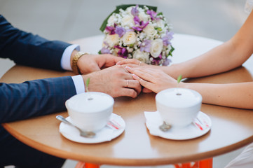 The bride and groom tenderly hold hands, sitting in a cafe at a wooden table with coffee. Photography, concept.