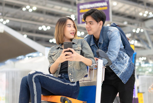 Romantic couple in airport. Young Asian couples are happy in the passenger Hall are ready for traveling while sitting on luggage trolley and using smartphone. Lover travel and transportation concept