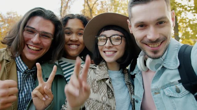 POV portrait of joyful men and women taking selfie in park showing thumbs-up smiling looking at camera. Modern technology and friendship concept.
