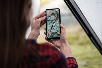 Women hands holding smartphone with app navigation hiking map on screen.  Mountains map with route and markers. Girl planning a trip inside the tent at the camp. 