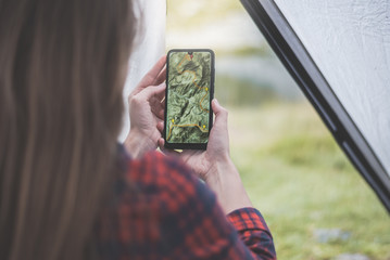 Women hands holding smartphone with app navigation hiking map on screen.  Mountains map with route and markers. Girl planning a trip inside the tent at the camp. 
