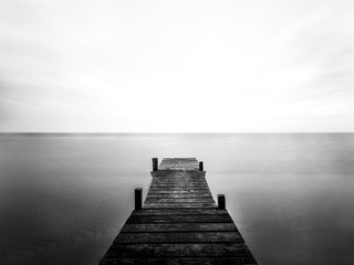 Greyscale of a wooden bridge on the sea under the sunlight and a cloudy sky