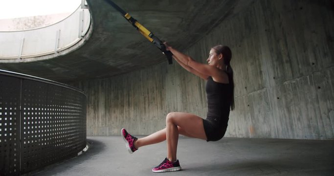 Low side shot of purposeful Caucasian female using TRX holding loops training alone, motivation of healthy body achieve intense girl exercising squatting stretching leg forward outdoor urban
