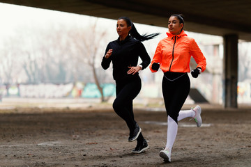 Two young fitness woman running under overpasses during cold day