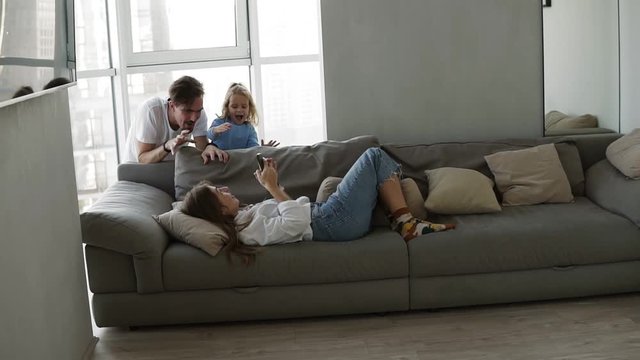 In living room tired mother laying on sofa with smartphone, adorable daughter and father suddenly appear from the back couch and scaring mother. Couple and child having fun together at home, enjoy