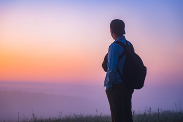 young man with a backpack watching the sunrise on nature. male tourist hiker looking at on a foggy field and mountains and enjoying breathtaking sunrise. Active life, adventure, travel concept.