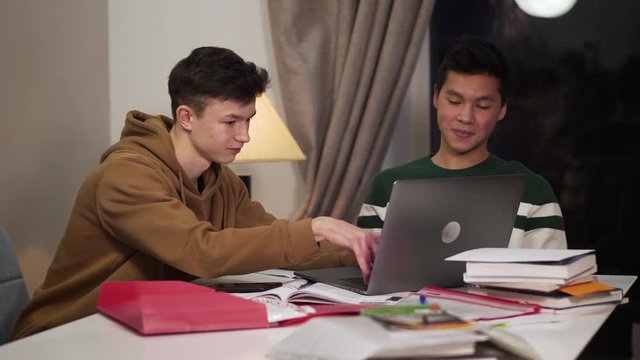 Two intelligent smiling college students sitting at the table and using laptop. Happy Caucasian and Asian teenagers studying online and giving high five. Happiness, intelligence, education.
