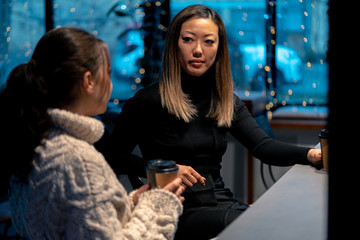 Asian Young Woman Attentively Listening Her Friend In Oversize Sweater And Drinking Coffee.