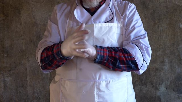 elderly man with gray beard in kitchen suit and white apron wears thin gloves before working with meat