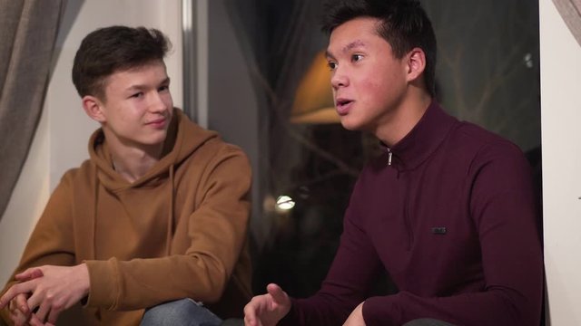 Portrait of Caucasian and Asian friends talking and giving dap. Teenage friends spending evening indoors. Happy people smiling. Friendship, support, lifestyle.