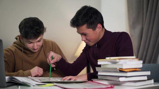 Close-up portrait of two excited boys looking at map and discussing geography. Smiling Caucasian and Asian college students talking and giving dap. Lifestyle, joy, education concept.