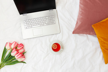 Open laptop, cup of tea with lemon, velvet pink and yellow pillows, bouquet of pink tulips flowers lying on bed with white linens. Work at home concept. Female workspace background. Flat lay, top view
