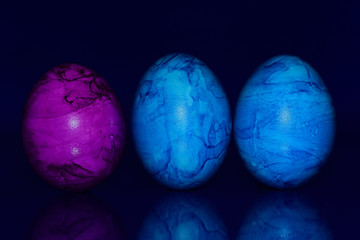 Three marbled, colored, blue and pink Easter eggs stand side by side against a reflective blue background and are reflected on the floor