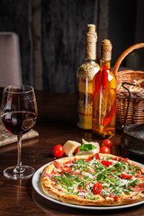 Italian food. Classic thin pizza with large sides, prosciutto, cherry tomatoes, arugula, parmesan cheese. Serving dishes in a restaurant on a white plate with red wine.