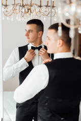 Preparations for the wedding. Wedding morning groom. Groom straightens the bow tie, handsome groom getting dressed and preparing for the wedding. The groom reflects in the mirror