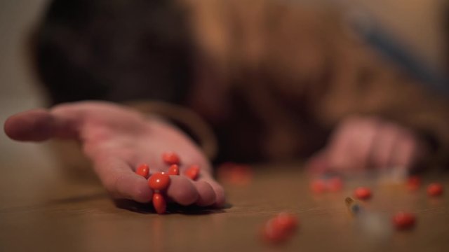 Close-up of orange drug pills falling out from male Caucasian hand, blurred boy lying on the floor at the background. Young narcomaniac having overdose. Drug dependence, risk, harm.