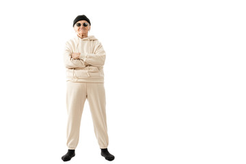 Naklejka premium Stylish old hipster woman in beige hoodie black hat and sunglasses stands with crossed hands and looking at the camera isolated over white background