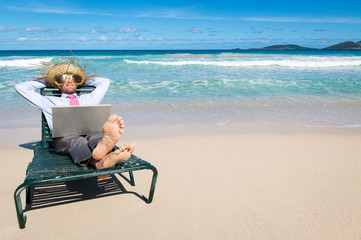 Barefoot businessman kicking back in a beach chair with laptop on his lap and sunglasses and straw...