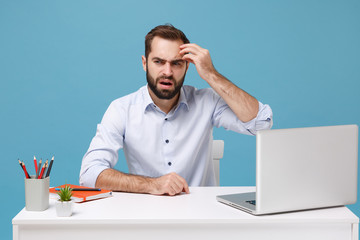 Bewildered young bearded man in light shirt sit and work at desk with pc laptop isolated on pastel...