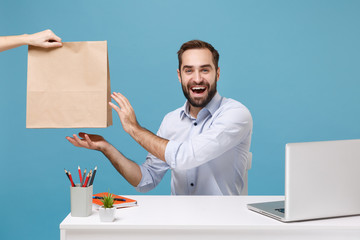 Funny man in shirt sit work at desk with pc laptop isolated on blue background. Food products delivery courier service from shop or restaurant to office Taking brown clear empty blank craft paper bag.