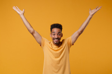 Smiling young african american guy in casual t-shirt posing isolated on yellow orange wall background, studio portrait. People emotions lifestyle concept. Mock up copy space. Rising spreading hands.